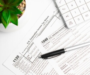 Tax forms 1040 and 1120 with a pen, calculator, and a potted plant on a white desk.