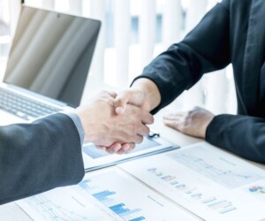 Two people in business attire shake hands over charts and a laptop on a desk in an office setting.