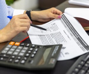 Person reviewing a document with a pen, beside a calculator and a keyboard on a desk.